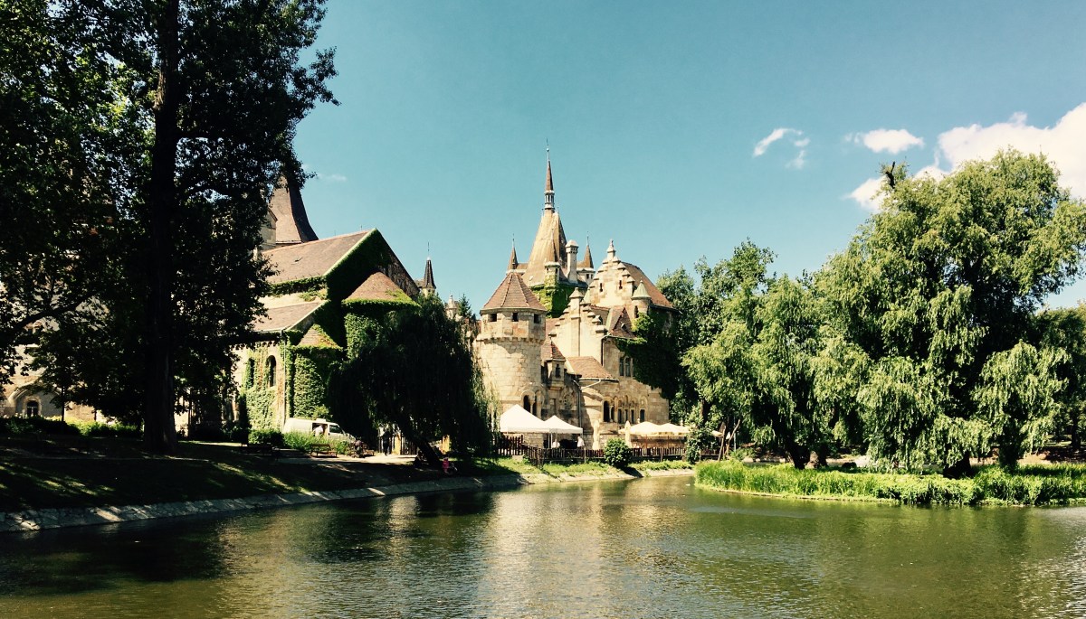 Image of a large sprawling building on the banks of a lake. There are plants climbing up the left building and trees on either side.