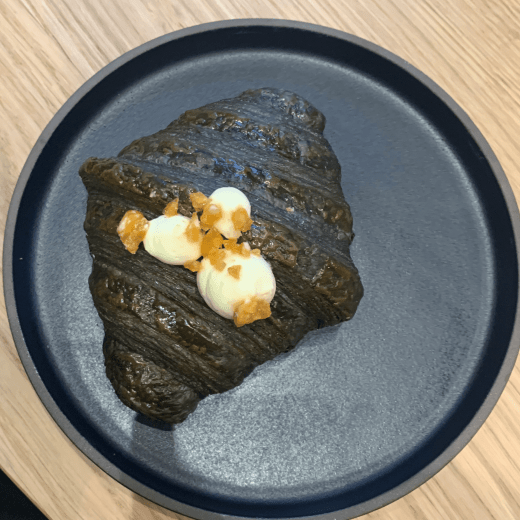 Black caramelised banana croissant with 3 blobs of cream on it sitting on a round metallic plate on a wooden table.